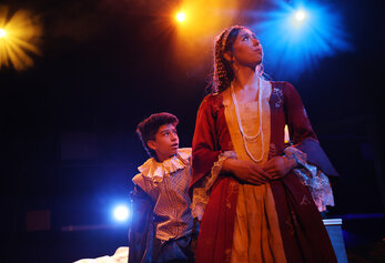 Two students in period costumes perform a dramatic scene in University High School’s “Romeo & Juliet,” lit by warm stage lighting.
