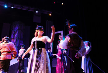 Students perform a masked dance scene in University High School’s “Romeo & Juliet,” wearing period costumes under colorful stage lighting.